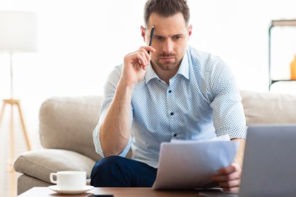 Household Financial Management Concept. Focused Man Reading Tax Or Debt Notification Papers Sitting At Laptop Indoor